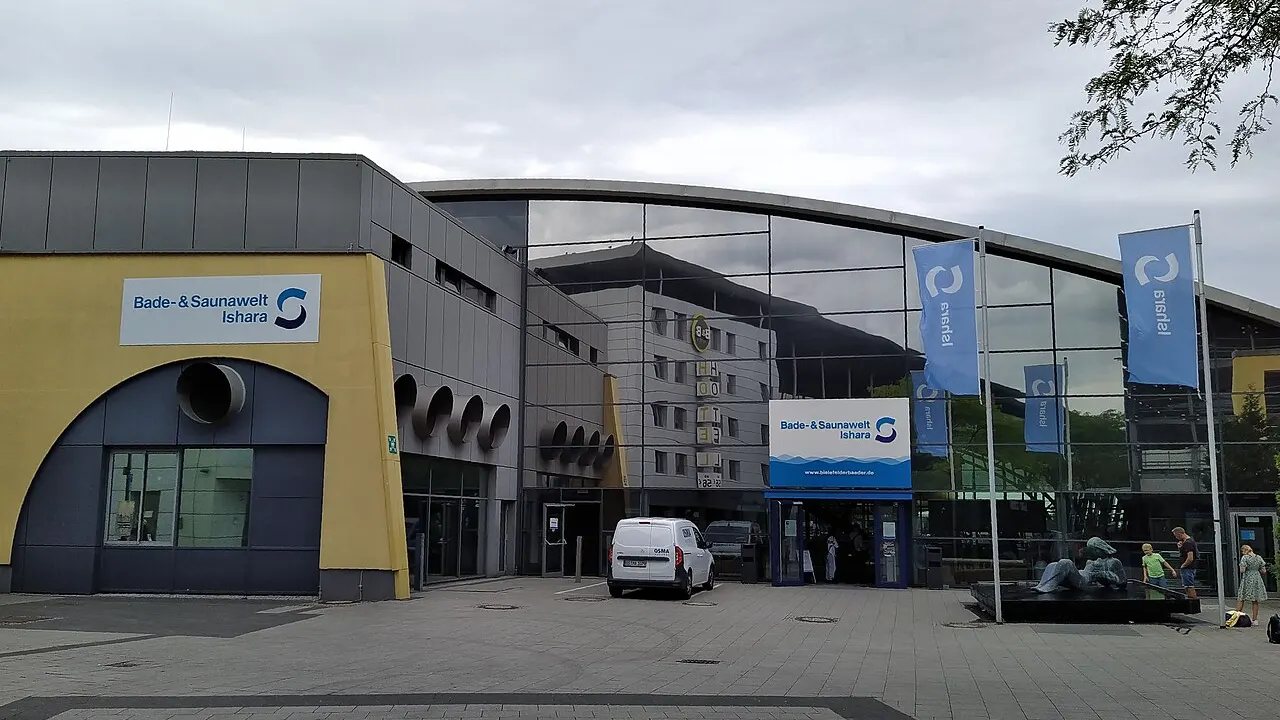 Eingang der Bade- und Saunawelt Ishara in Bielefeld mit Glasfassade, gelbem Gebäudeteil und blauen Ishara-Fahnen