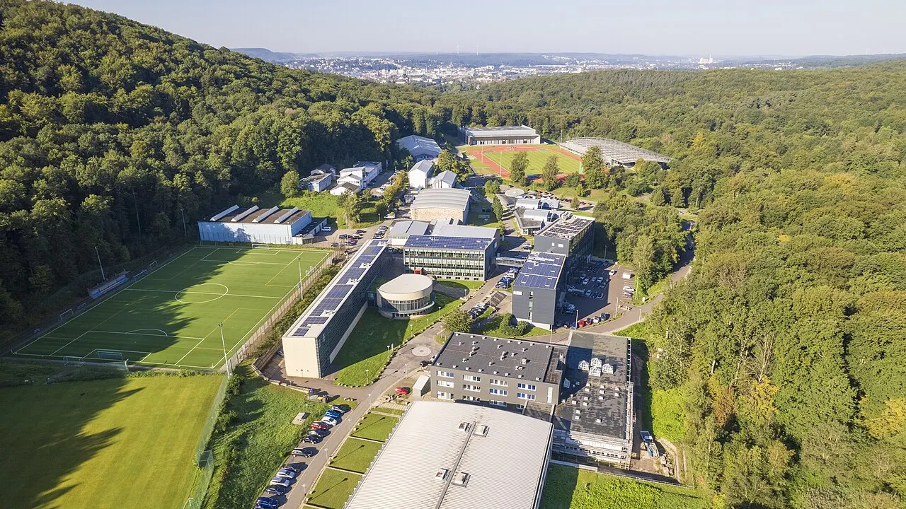 Luftbild des Sportcampus Saar mit Sporthallen, Fußballplatz und Wald, im Hintergrund die Stadt Saarbrücken