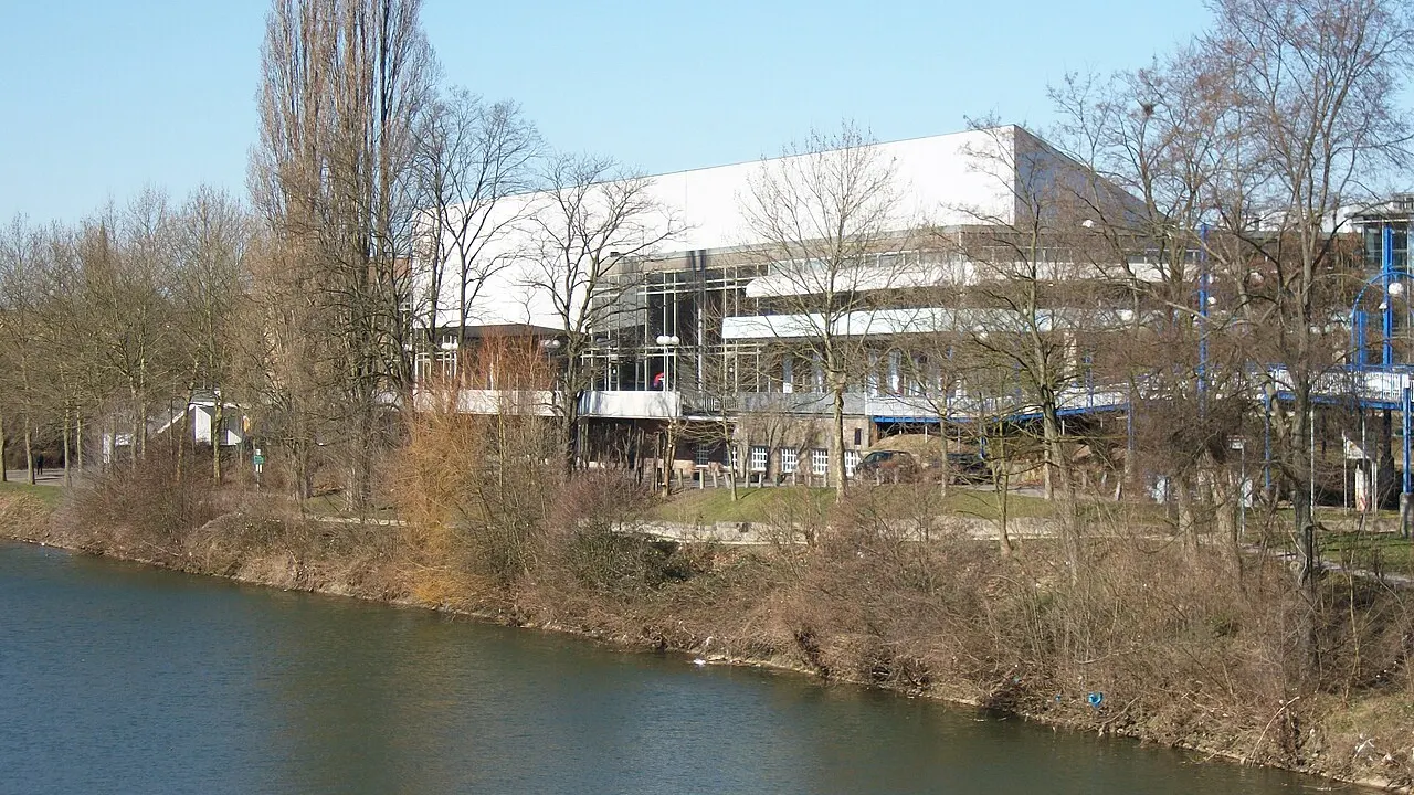 Die helle Congresshalle Saarbrücken hinter kahlen Bäumen am Ufer der Saar unter blauem Himmel