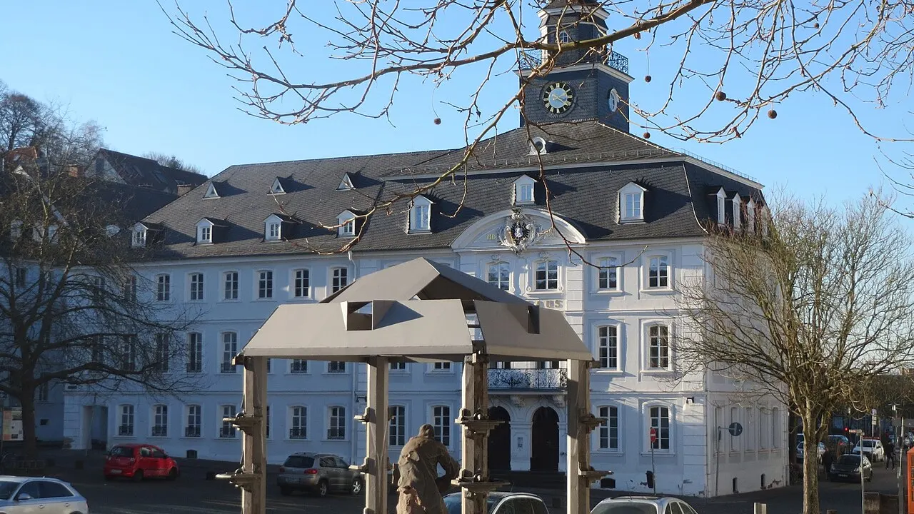 Das weiße Alte Rathaus mit Uhrturm am Schlossplatz in Saarbrücken hinter kahlen Bäumen, davor ein Brunnen mit Bronzefigur